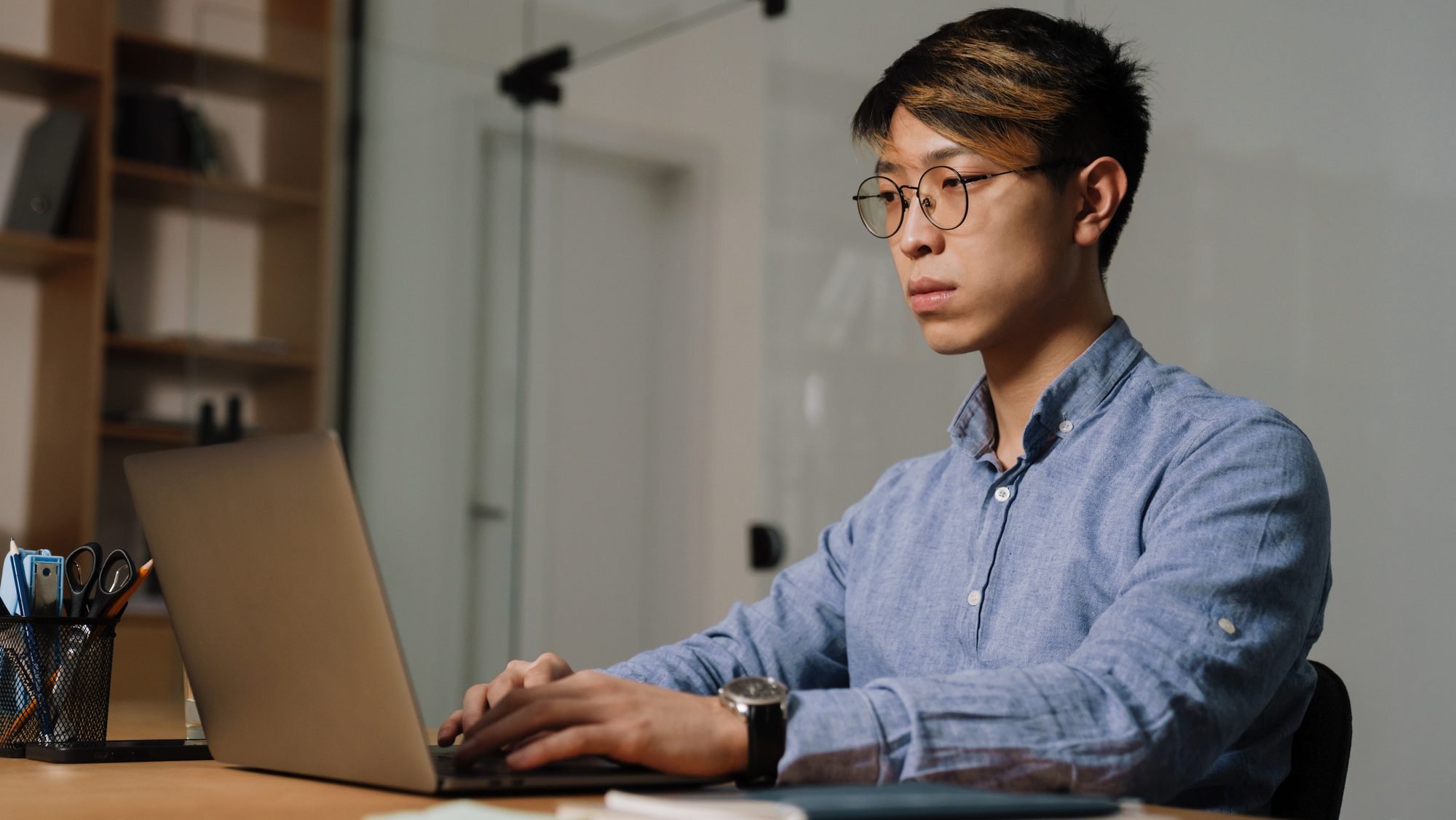 Asian guy in eyeglasses working with laptop while sitting at table in office.
