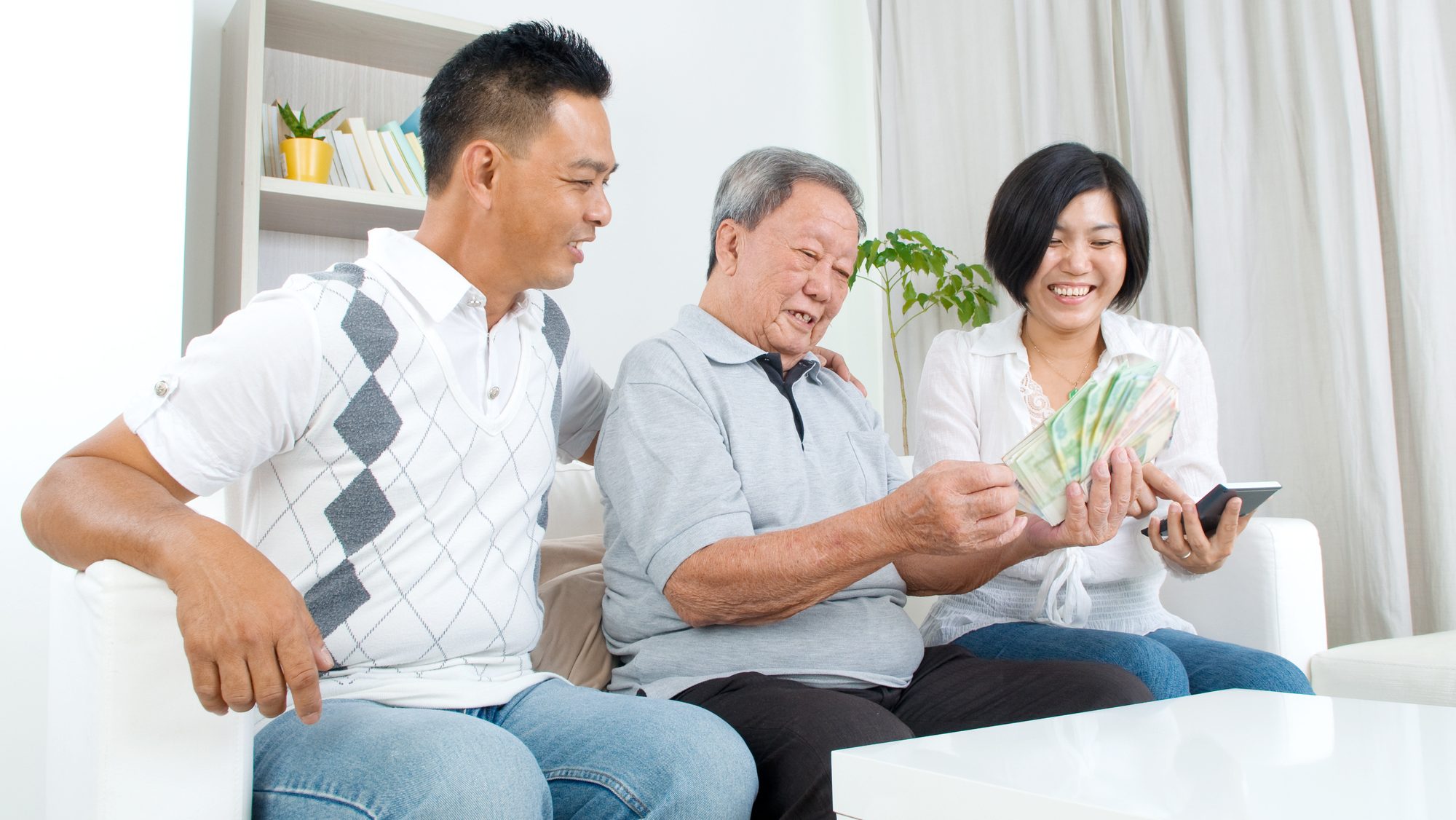 Asian family sitting on a sofa with Dad in the middle holding a fan of cash.