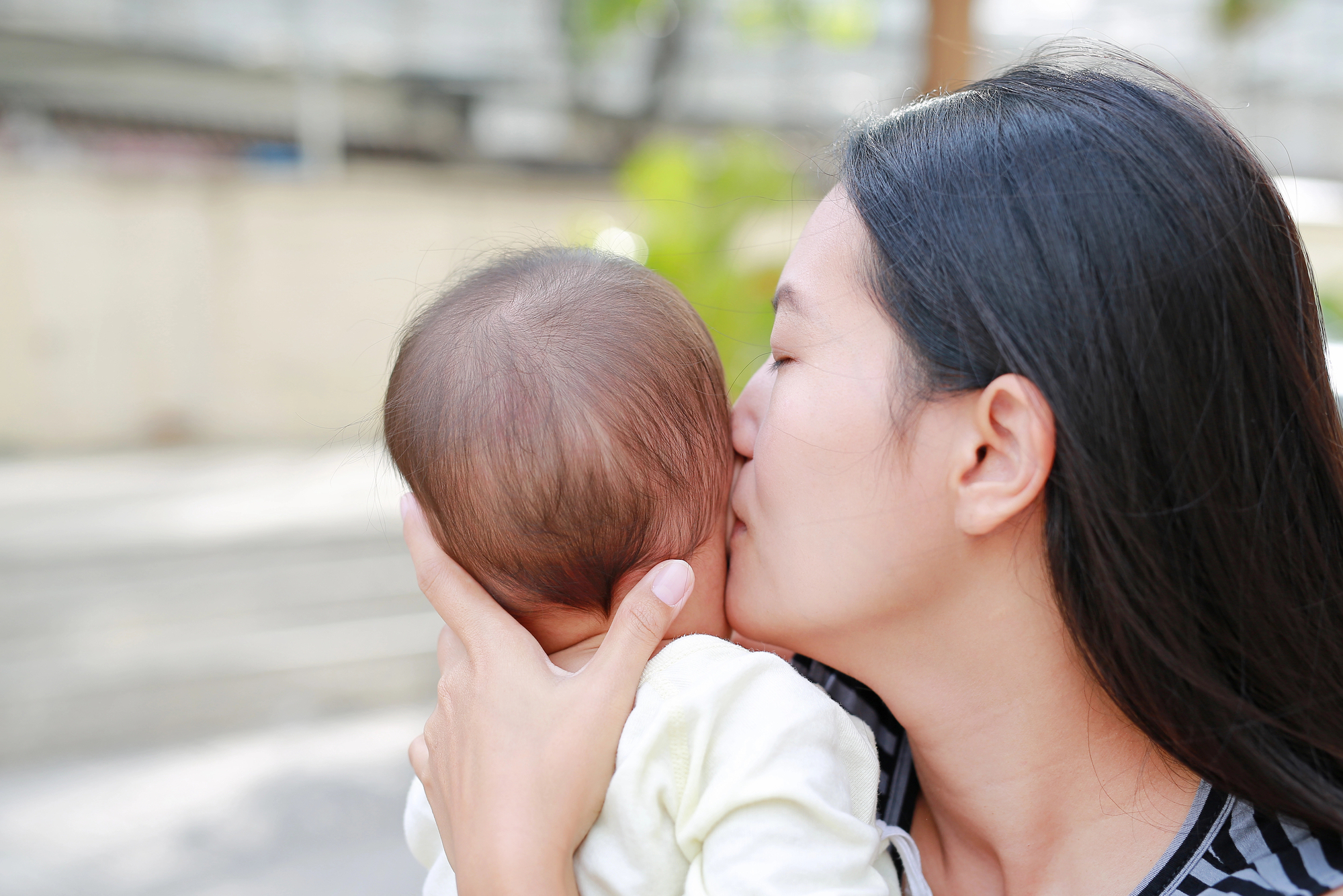 Asian mother carrying and kissing her infant outdoors.